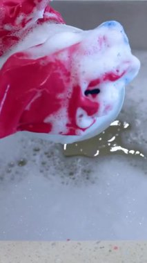 Hands in red gloves wash a dish with a soapy sponge in a kitchen sink