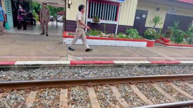 A bustling train station in Kanchanaburi, Thailand, with people in uniform and travelers. Bright daylight enhances the lively atmosphere
