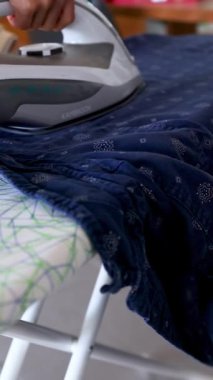 Bir kişi ütü masasında koyu mavi bir giysi ütüler, iyi aydınlatılmış bir odada pürüzsüz, hassas hareketler sergiler.