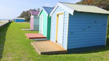 Canlı sahil kulübeleri parlak gün ışığında çimenli bir yol çiziyor, sakin bir sahil atmosferi sergiliyor.