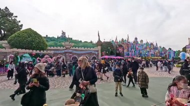 Ziyaretçiler Tokyo Disneyland 'de geziniyor, renkli mimarisi ve bulutlu gökyüzü altında hareketli kalabalıklar ile canlı bir atmosfer yakalıyorlar.