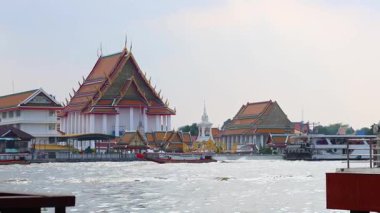 Uzun kuyruklu bir tekne Chao Phraya Nehri 'nde yol alır. Arkasında geleneksel Tayland mimarisi vardır.