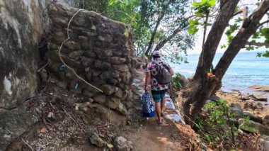 Phuket, Tayland 'da, verimli yeşillik ve berrak sularla çevrili, parlak gün ışığında bir insan okyanus kıyısında kayalık bir yolda yürür.