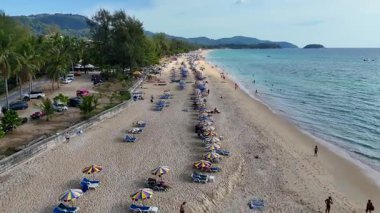 İnsansız hava aracı görüntüleri Phuket sahilini turistler, şemsiyeler ve açık mavi sularla aydınlık gün ışığında yakalıyor.