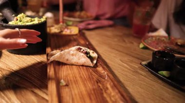 Bir el, Meksika yemekleri ile çevrili, sıcak bir aydınlatma altında, ahşap bir masada taco için uzanır.