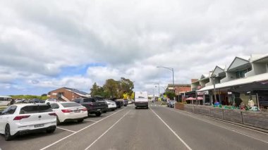 Büyük Okyanus Yolu 'ndaki hareketli bir kasabadan geçmek, bulutlu bir gökyüzünün altında park halindeki arabaları, dükkanları ve yayaları sergilemek.