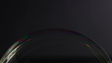 Bir sabun baloncuğu, dinamik ışık oyunu ve yüzey gerilimi gösteren karanlık bir arkaplan karşısında canlı renk yansımaları gösterir.