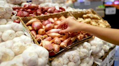 Bir el, sarımsak ve soğanla çevrili iyi aydınlatılmış bir süpermarkette sergilenen soğanlardan soğan seçer.