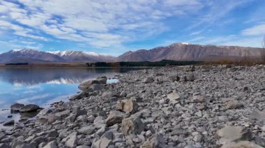 Hava görüntüleri Tekapo Gölü 'nün kayalık kıyı şeridini parlak mavi bir gökyüzünün altında net yansımaları ve dağlık bir zemin ile yakalıyor.