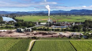 Drone footage captures a sugar mill surrounded by lush fields and mountains under clear skies in Murwillumbah, NSW