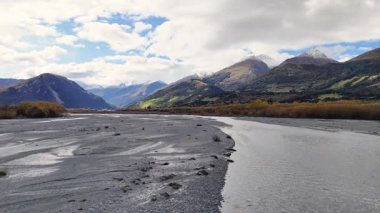 Glenorchy 'nin dağlık arazisi boyunca uzanan sakin bir nehir parçalı bulutlu bir gökyüzünün altında doğal gün ışığında yakalanır.