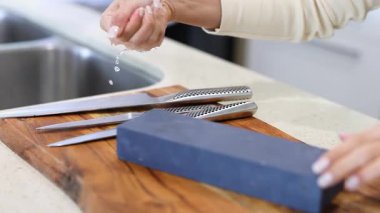 Bir insan, iyi aydınlatılmış bir mutfakta, ahşap bir tahtanın üzerinde bileme taşı kullanarak bıçakları biler.