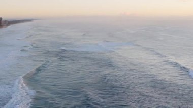 Yumuşak gündoğumu ışığı altında yumuşak okyanus dalgalarının havadan görünüşü sakin bir sahil manzarası yaratıyor.