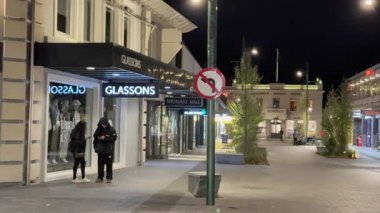 İki kişi gece Queenstown 'da, modern mağazalar ve şehir mimarisi ile çevrili, aydınlık, boş bir caddede yürüyor.