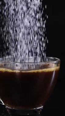 Şeker, koyu arkaplana karşı bir bardak çaya dökülüyor, granülleri ve sıvı etkileşimini vurguluyor.