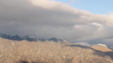 Clouds move swiftly over a rugged mountain range, casting dynamic shadows and highlighting the landscape's natural beauty