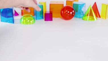 A hand interacts with colorful 3D geometric shapes on a white table, highlighting various forms and colors