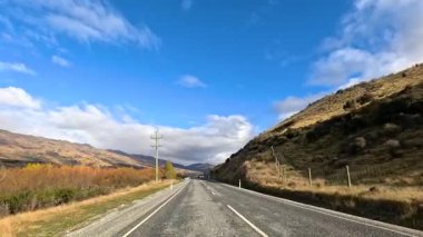 A vehicle travels a scenic road in Wanaka, New Zealand, surrounded by hills and clear skies, captured in bright daylight