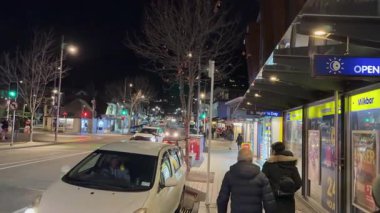 Two people walk past a brightly lit street in Queenstown at night, surrounded by shops and passing cars