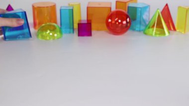 Hands interact with vibrant geometric shapes on a white table under bright lighting, demonstrating movement and arrangement