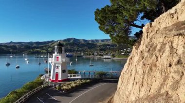 Aerial footage captures Akaroa's lighthouse and harbor under clear skies, showcasing serene waters and anchored boats