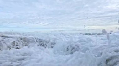 Dinamik okyanus dalgaları bulutlu bir gökyüzü altında kayalık kıyı şeridine çarpar dramatik sıçramalar ve köpükler yaratır.