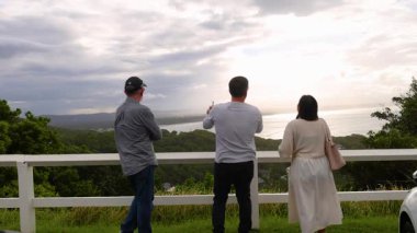 Byron Körfezi 'nde üç kişi huzurlu manzaranın tadını çıkarıyor. Bulutlu gökyüzü ve yemyeşil sakin, yansıtıcı bir atmosfer yaratır.