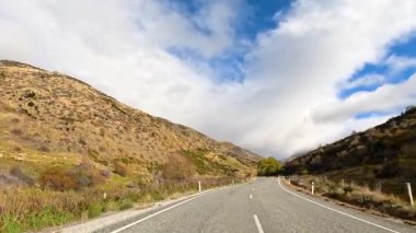 Bir araba Wanaka 'nın dolambaçlı yollarında, dağlık araziyle çevrili, dağınık bulutlu, canlı mavi bir gökyüzü altında seyahat ediyor.