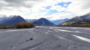 Dağları ve ağaçları olan sakin bir nehir yatağı parçalı bulutlu bir gökyüzünün altında doğal güzellik ve sakinlik sergiliyor.