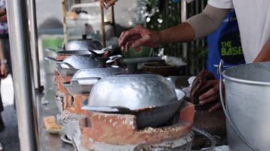 Eller, kalabalık bir Phuket pazarında sıcak tava kullanarak geleneksel Tayland atıştırmalıklarını ustalıkla hazırlar. Doğal ışıklandırma hareketli sokak sahnesini vurguluyor