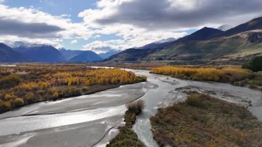 Glenorchy, Yeni Zelanda 'da dinamik bir gökyüzü altında hareketli sonbahar yaprakları ve görkemli dağlar arasında sakin bir nehir kıvrılır.