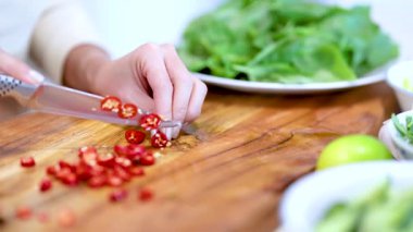 Ahşap bir tahtanın üzerinde kırmızı biber dilimleyen eller. Yanında taze yeşillikler ve limonlar var. Parlak ışık altında.