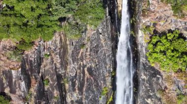 Drone captures a stunning vertical waterfall cascading through dense greenery and rocky cliffs in a vibrant rainforest setting