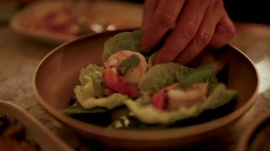 Bir el, karides ve yeşillikleri yumuşak, sıcak aydınlatmanın altında bir tabağa yerleştirir ve davetkar bir mutfak sahnesi yaratır.