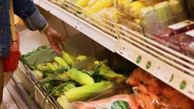 Parlak ışık altında süpermarketten mısır toplayan bir insan taze ürün seçimi sergiliyor.