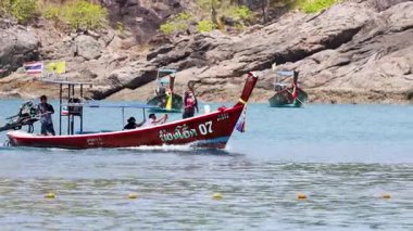 Uzun kuyruklu bir tekne, Phuket 'in sakin sularında gezinerek, kayalık bir kıyı şeridinde yerel balıkçılar ve turistleri gösteriyor.