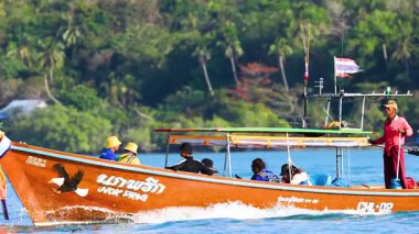 Uzun kuyruklu bir tekne Phuket, Tayland 'ın dingin sularında yol alır. Parlak gün ışığında, arka planda yemyeşil bir alan vardır.