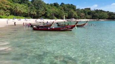Phuket, Tayland 'da canlı bir sahilin havadan görünüşü. Parlak güneş ışığı altında uzun kuyruklu tekneler, yüzücüler ve yemyeşil alanlar.