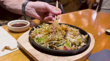 Çubuklar, canlı sebzelerle çevrili ve sıcak bir ortamda sake eşliğinde cızırdayan bir tabaktan buharlı şehriyeleri kaldırır.