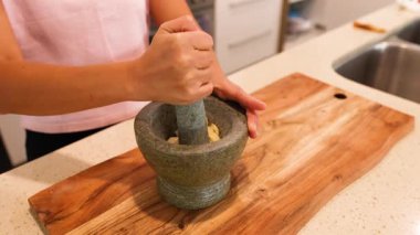 Bir insan, iyi aydınlatılmış bir mutfakta ahşap bir tahtanın üzerinde havan topu ve tokmakla malzemeleri öğütür.