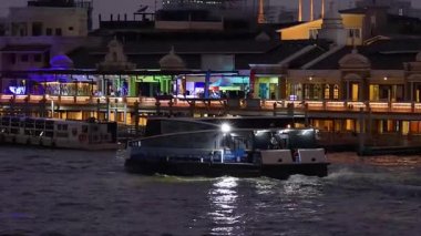 Tekne nehir kenarındaki aydınlık binaların önünden geçiyor.