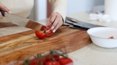 Parlak bir mutfakta, ahşap bir tahtada domatesleri dilimleyip beyaz bir kaseye koyan bir insan.