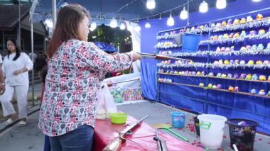 Canlı bir Bangkok tapınağı fuarında, parlak ışıklar ve renkli bir çevre altında bir kadın hava tabancası atışına katılıyor.