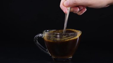 Bir el, cam bardakta koyu bir fonda kahve karıştırıyor, dönen hareketi ve zengin rengi vurguluyor.