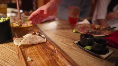 Bir el, yemek ve içeceklerle çevrili sıcacık bir Meksika restoranında, ahşap bir masanın üzerindeki taco 'ya uzanır.