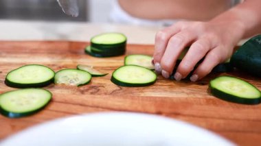 Parlak bir mutfakta ahşap bir tahtanın üzerinde salatalık dilimleyen eller. Yakın çekim, doğal ışıklandırma, taze ürünlere odaklan