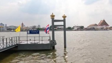Bangkok 'ta bulutlu gökyüzünün altında bir rıhtım, bayraklar ve uzak tapınaklarla sakin bir nehir manzarası.