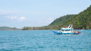 Tayland 'ın Phuket kentindeki yemyeşil tepelerin yakınlarındaki berrak mavi sularda gün ışığında bir tekne seyahat eder.