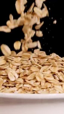 Yulaf taneleri bir kase içinde siyah bir arkaplana doğru taşar. Ağır çekimde dramatik bir ışık altında yakalanır.