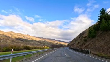 Wanaka 'nın dolambaçlı yollarında sakin bir sürüş, parlak mavi gökyüzünün altında çarpıcı dağ manzaraları sergiliyor.